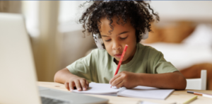 Child with headphones sitting and writing at a desk in front of a computer.